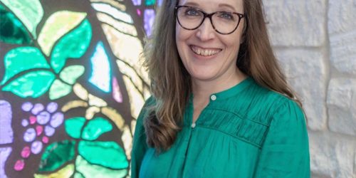 erin-swenson-reinhold_500px500px A woman in a green shirt stands in front of a green and purple stained glass window smiling.