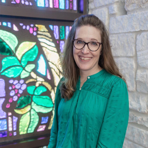A woman in a green shirt stands in front of a green and purple stained glass window smiling.