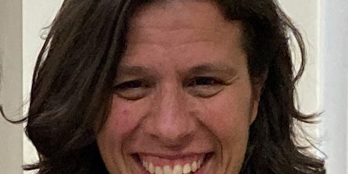 A woman with a white collar of a pastor stands in front of a beige wall smiling warmly.