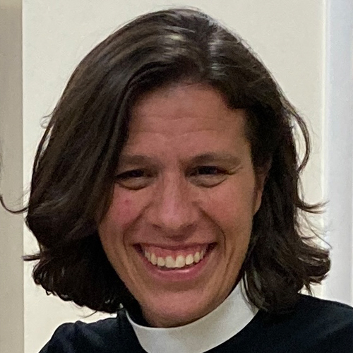 A woman with a white collar of a pastor stands in front of a beige wall smiling warmly.