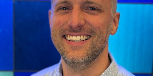 A smiling man in a light patterned collared shirt stands in front of a blue stained glass background of different shades of squares.