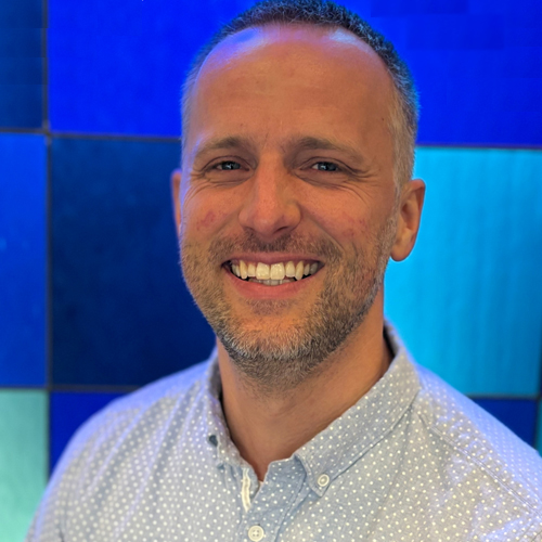 A smiling man in a light patterned collared shirt stands in front of a blue stained glass background of different shades of squares.