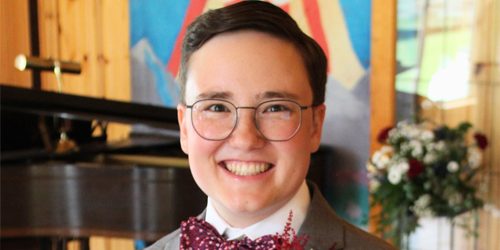 A young person smiles warmly wearing a bow tie and floral decoration in the lapel of their suit jacket in front of a piano and large painting of a dove rising.