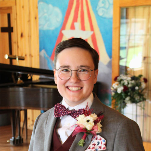 A young person smiles warmly wearing a bow tie and floral decoration in the lapel of their suit jacket in front of a piano and large painting of a dove rising.