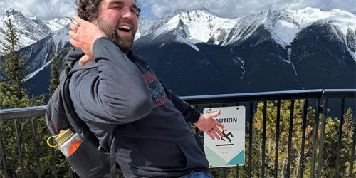 wes_kimball_500px500px_ A man kicks his leg out with in a fun manner at a mountaintop lookout, wearing a backpack and casual clothes.