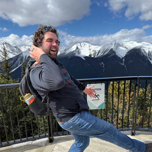 A man kicks his leg out with in a fun manner at a mountaintop lookout, wearing a backpack and casual clothes.